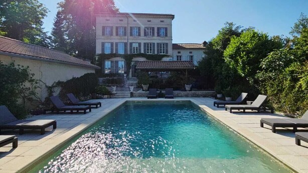 A rectangular swimming pool with lounge chairs on both sides, surrounded by a building with a tiled roof and a garden area. - The Paris Gîte (Ribérac)