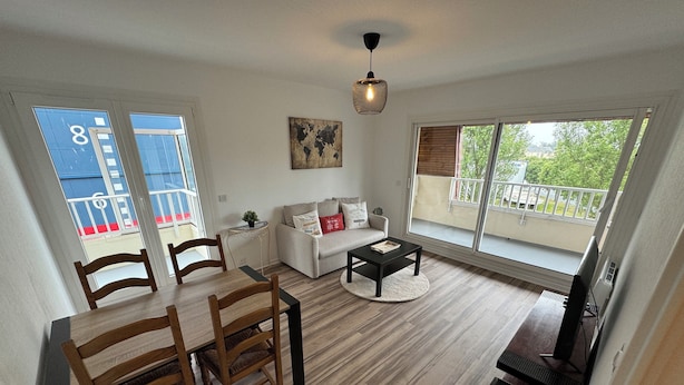 A living room with a dining area, a sofa, a coffee table, and a TV. There is a balcony with a railing and a view of the outside. - Large modern F3 - near port (Courseulles-sur-Mer)