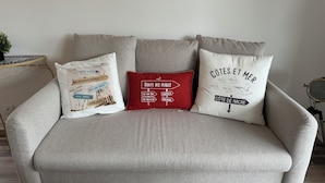 A grey sofa with three decorative pillows, one with a map design, one with a red and white sign, and one with text and birds. - Large modern F3 - near port (Courseulles-sur-Mer)