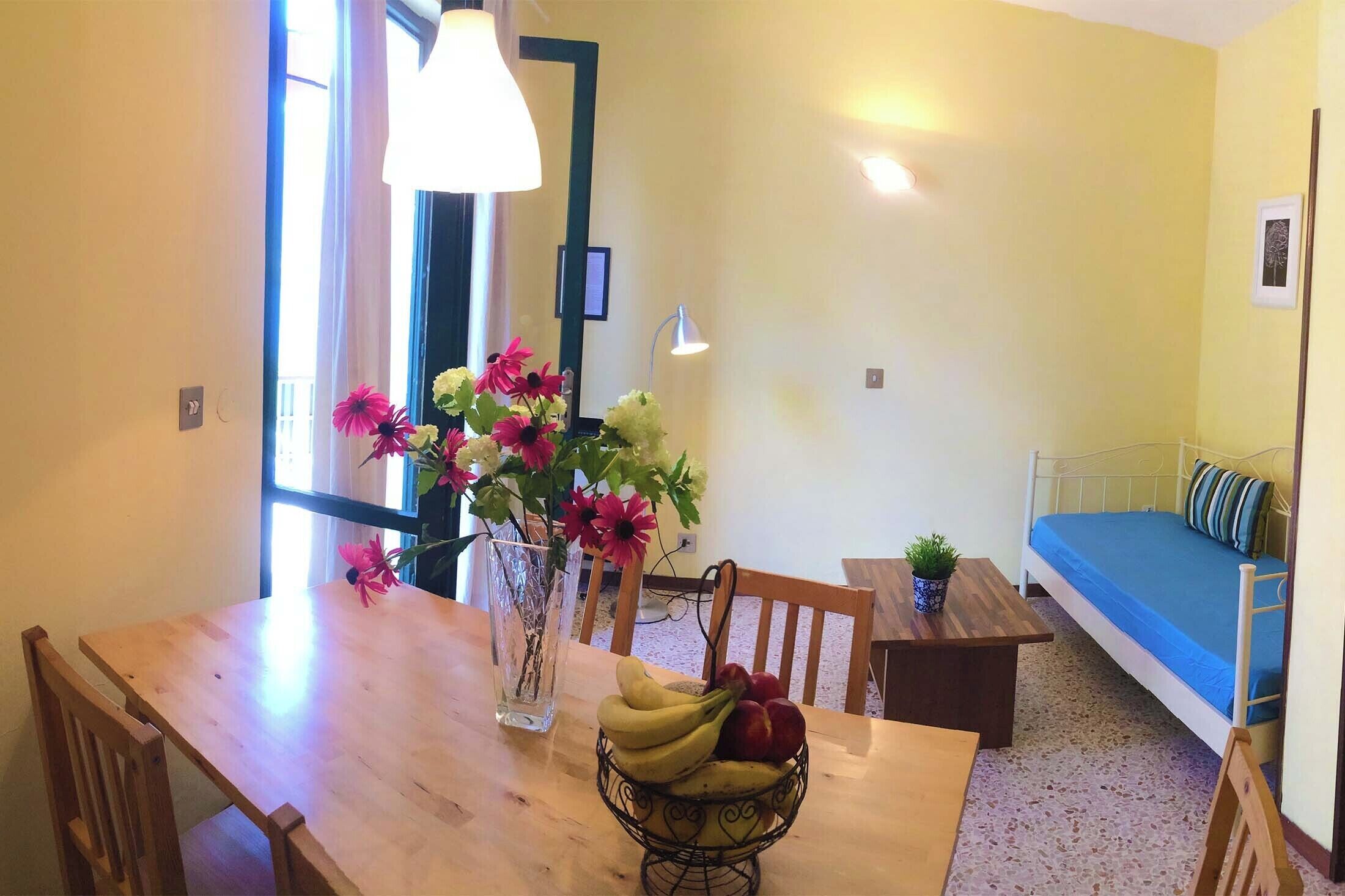 A dining area with a wooden table, chairs, a vase with flowers, and a fruit bowl.