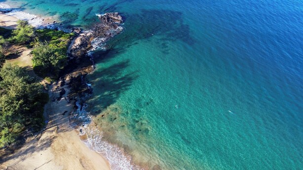 An aerial view of a coastal area with clear turquoise waters, sandy beach, and rocky outcrops.