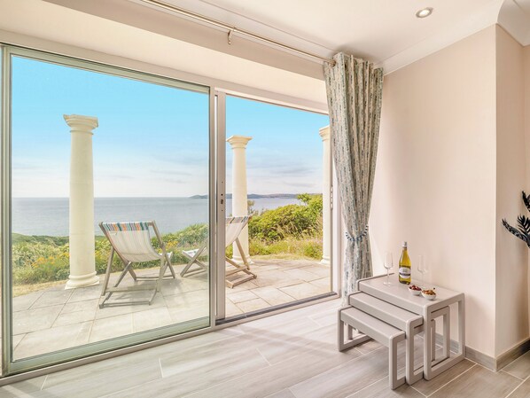 A balcony with a view of the sea, two chairs, a table with a bottle and glasses, and a curtain.