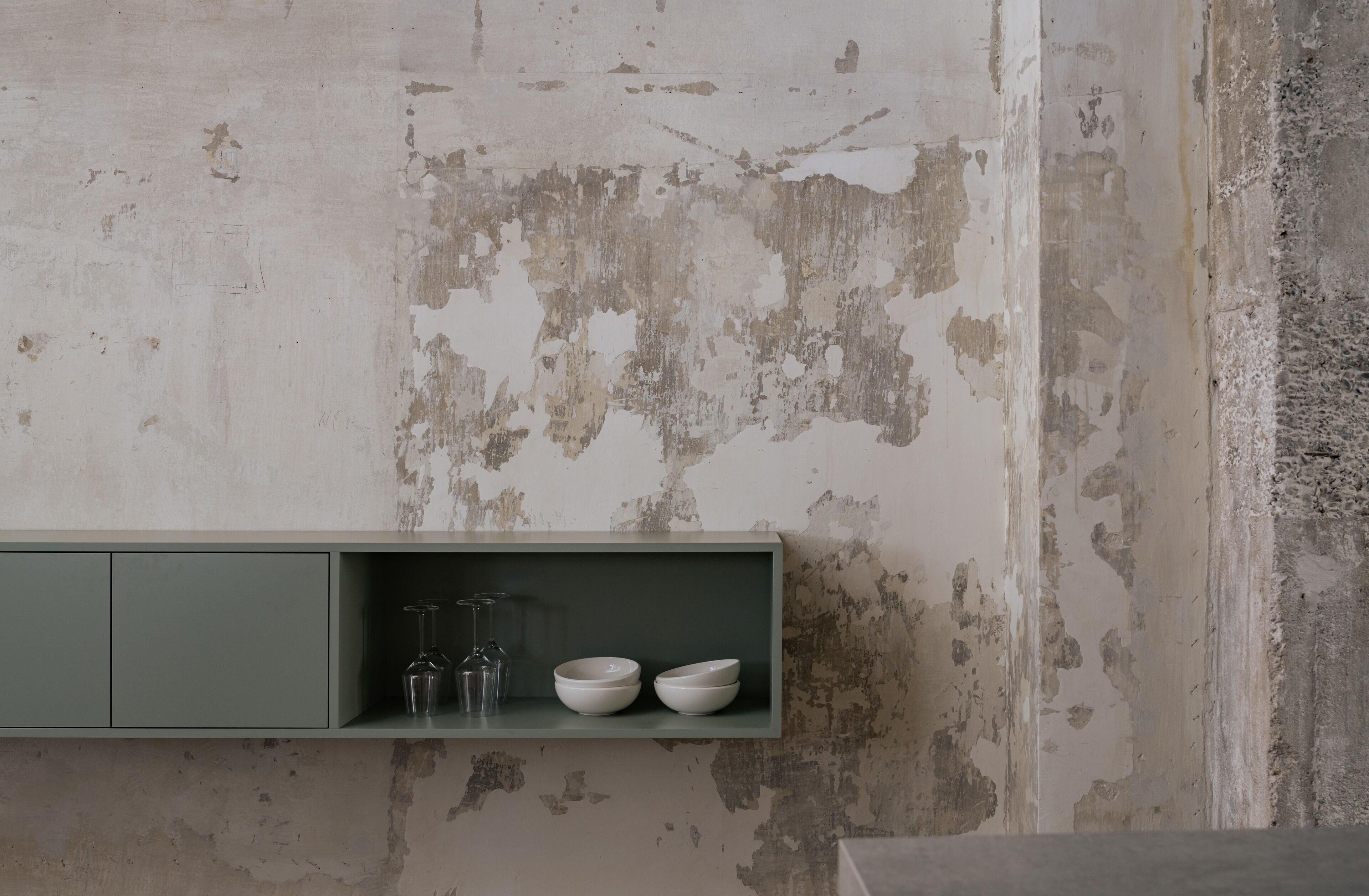 Cocina minimalista con pared de hormigón, un mueble verde y dos cuencos blancos sobre una repisa.