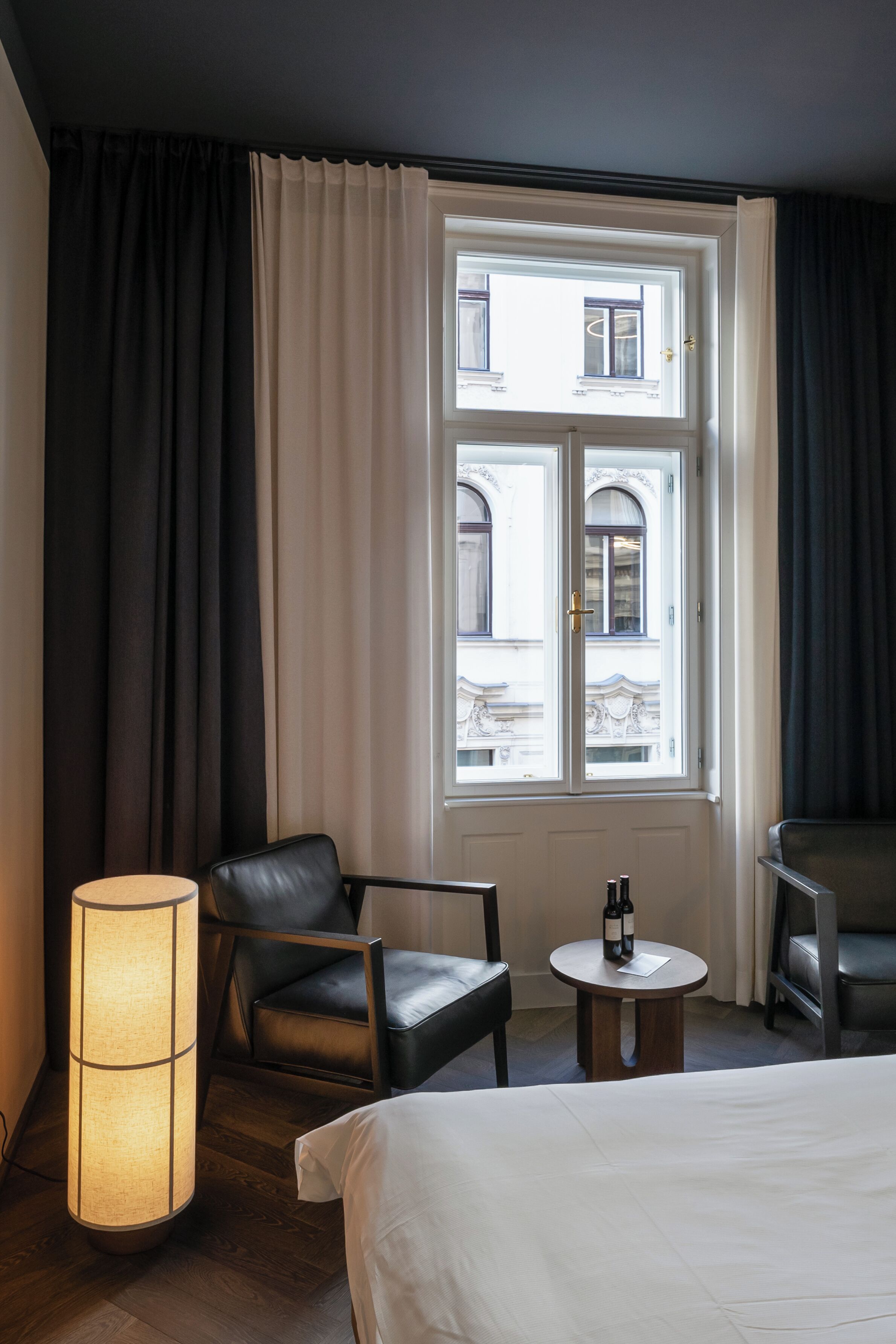 Habitación de hotel con cama, una silla, una mesa con una botella, lámpara y ventana con cortinas.
