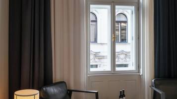 Habitación de hotel con cama, una silla, una mesa con una botella, lámpara y ventana con cortinas.
