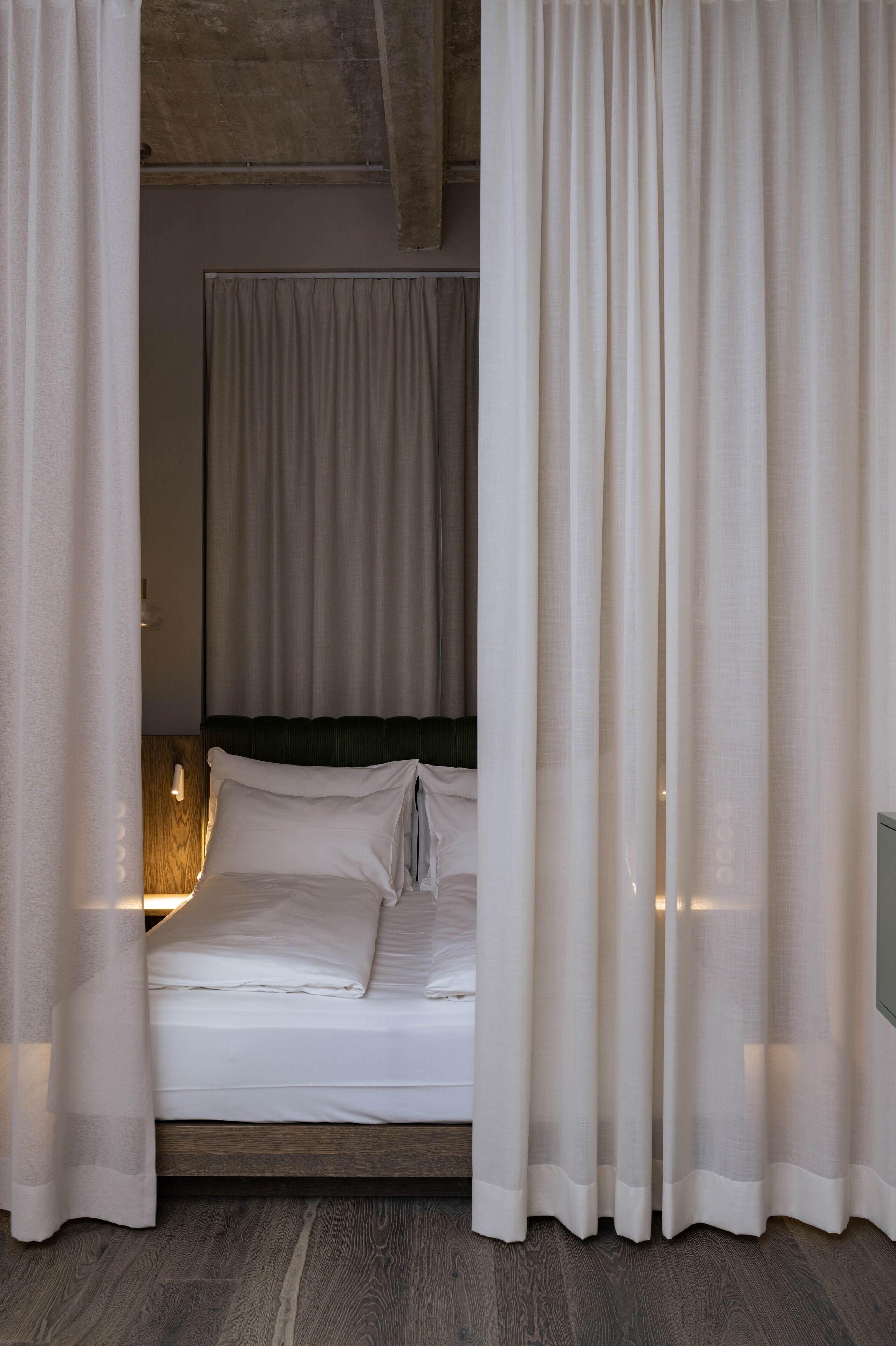 A neatly made bed with white linens and a wooden headboard, curtains partially drawn, and a wall-mounted light fixture.