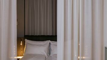 A neatly made bed with white linens and a wooden headboard, curtains partially drawn, and a wall-mounted light fixture.