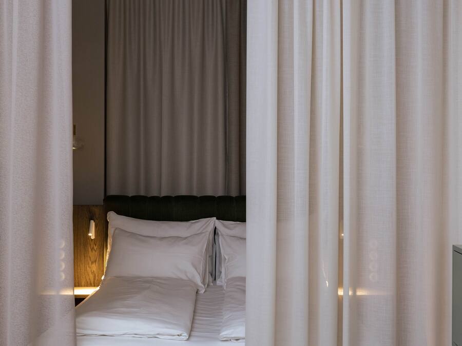 A neatly made bed with white linens and a wooden headboard, curtains partially drawn, and a wall-mounted light fixture.