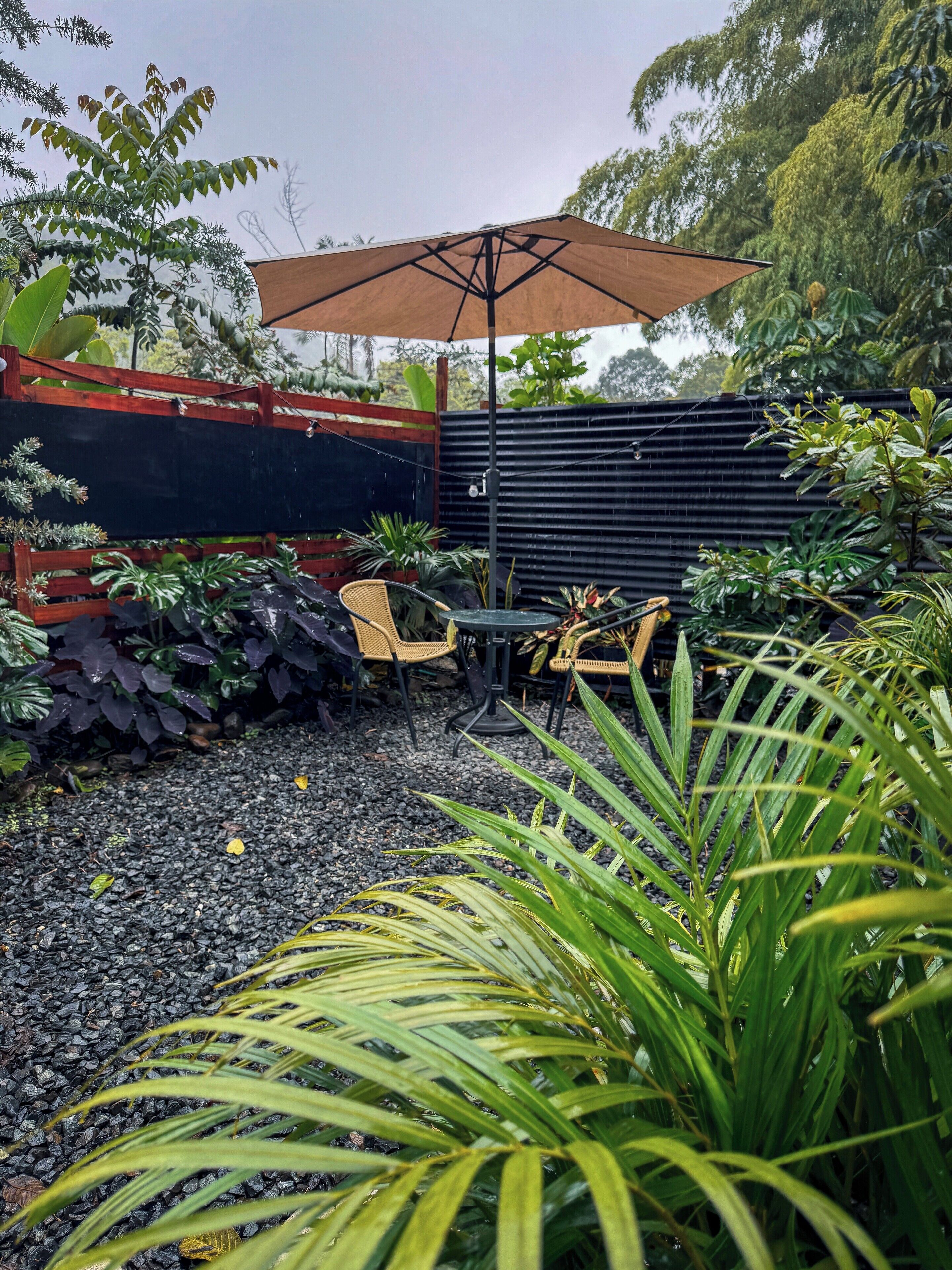 Un patio al aire libre con mesa y sillas bajo un amplio paraguas, rodeado de abundante vegetación y una valla de metal negro.