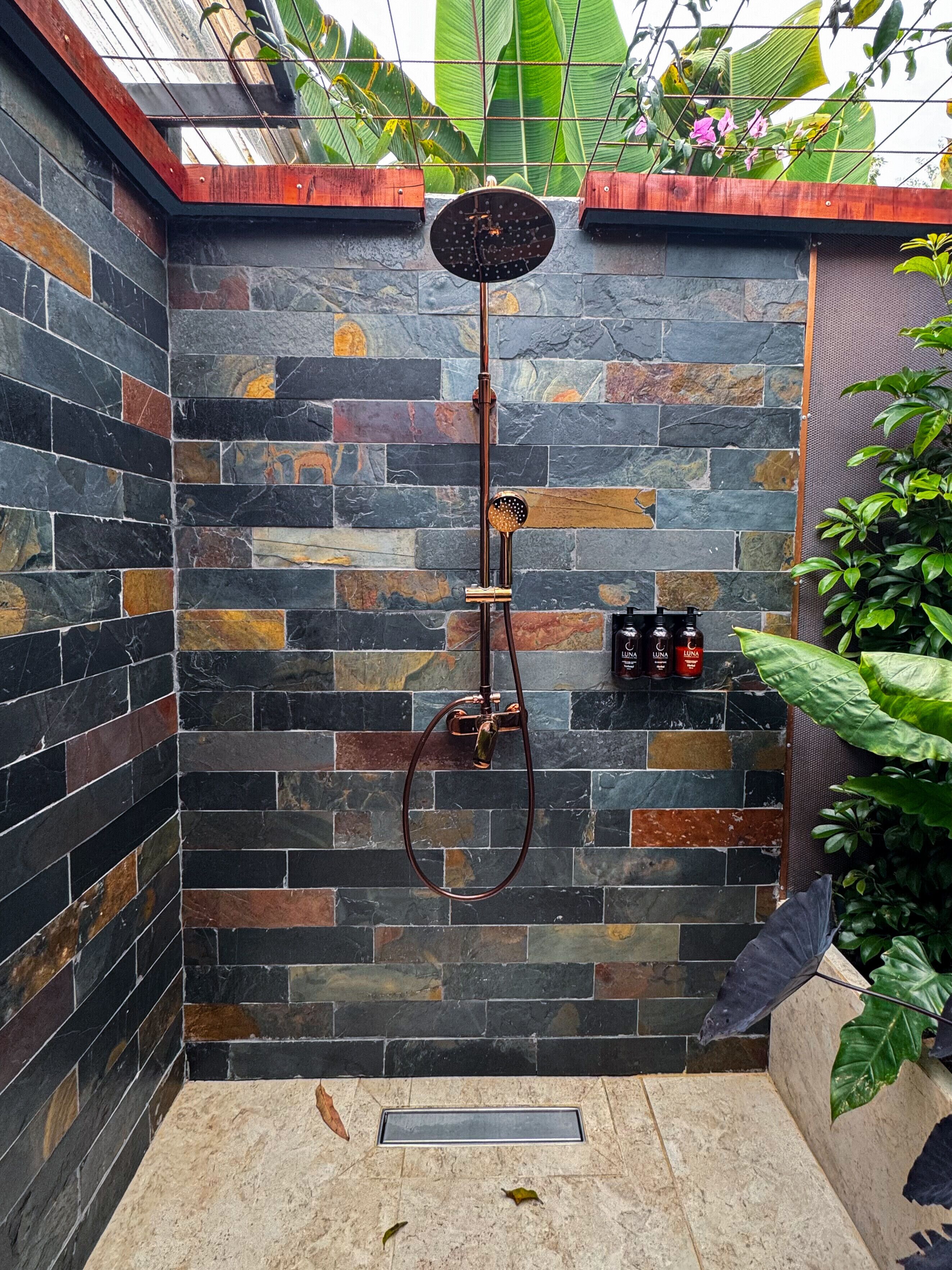 Zona de ducha con pared de piedra, ducha de lluvia y un estante pequeño con botellas.