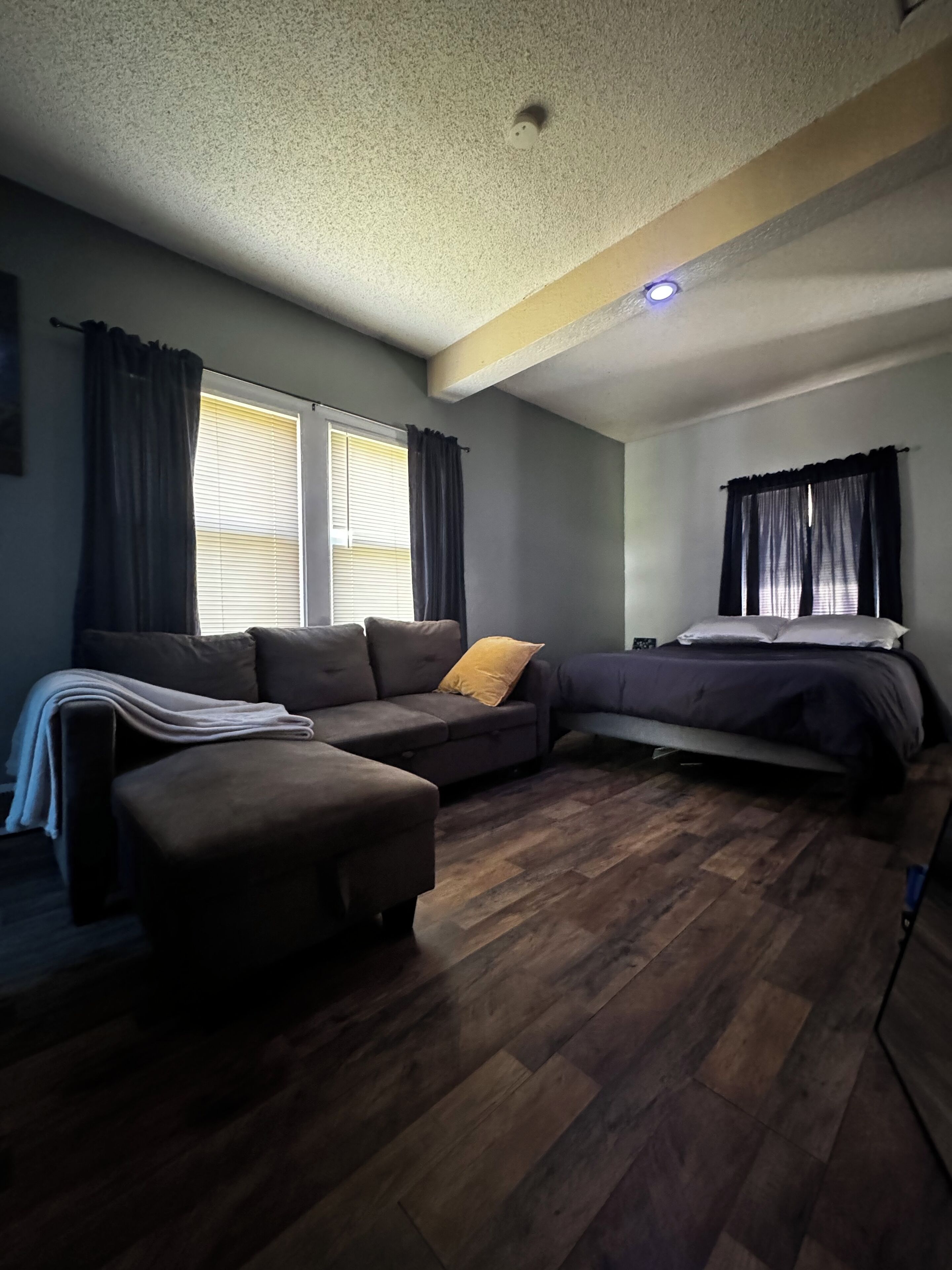 A room with a sectional sofa, a bed, a small table, and a window with curtains.