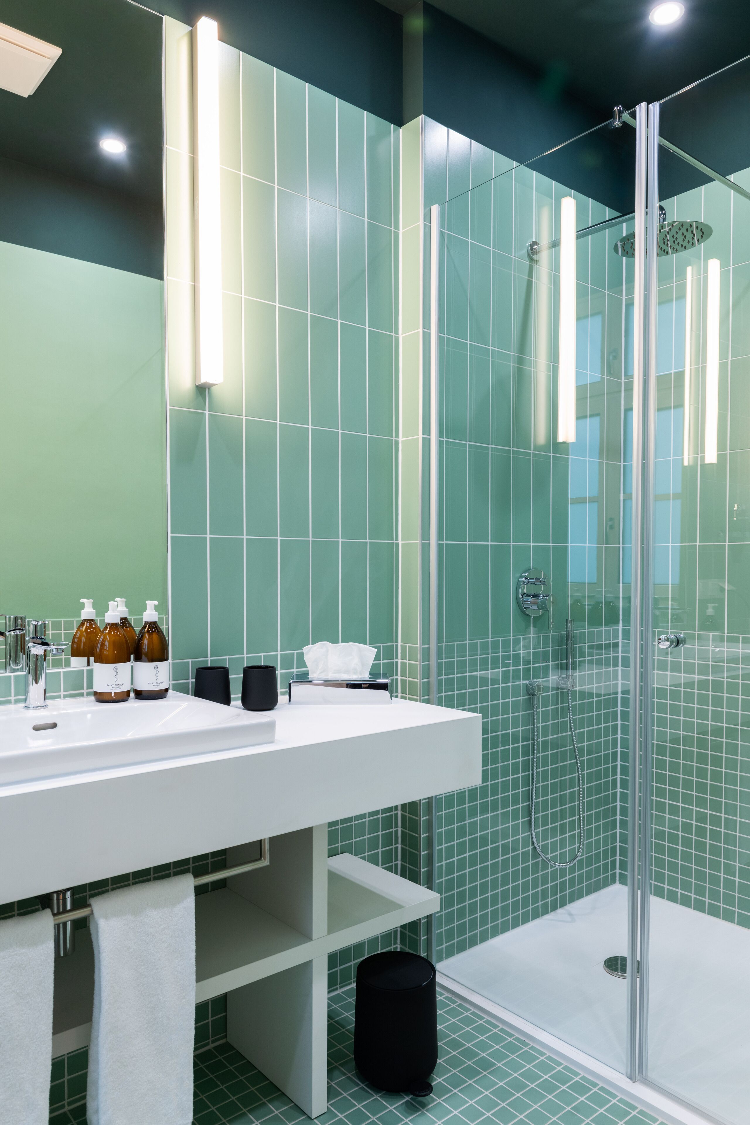 Une salle de bain moderne avec une douche à parois de verre, un lavabo blanc agrémenté d’articles de toilette et un mur recouvert de carreaux verts.