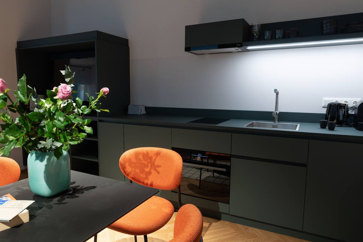 A modern kitchen with a dark countertop, a sink, and a built-in oven. There is a vase with flowers on the table and a chair with a floral design.