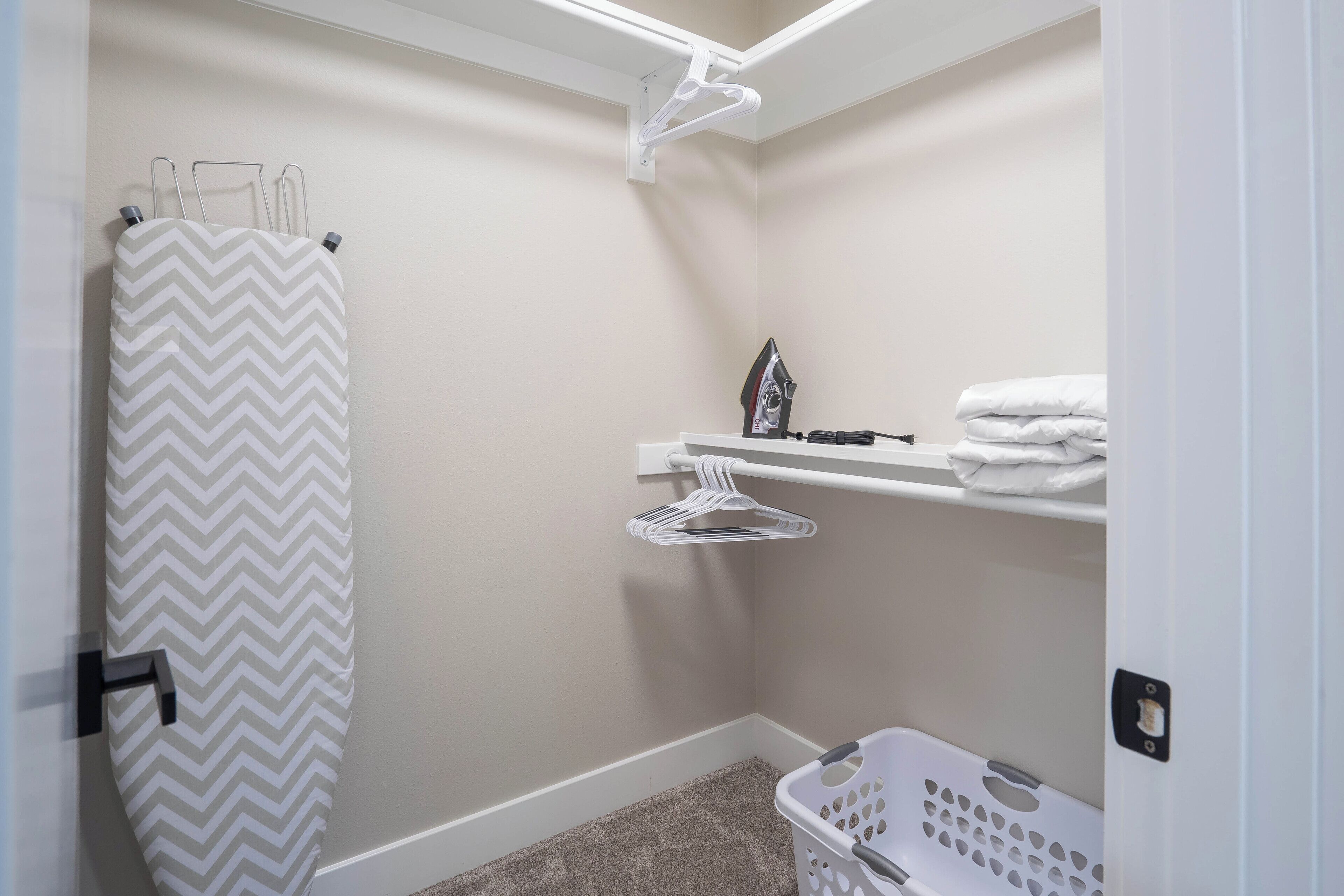 A laundry room with a chevron-patterned shower curtain, a white laundry basket, and a shelf with folded towels and an iron.