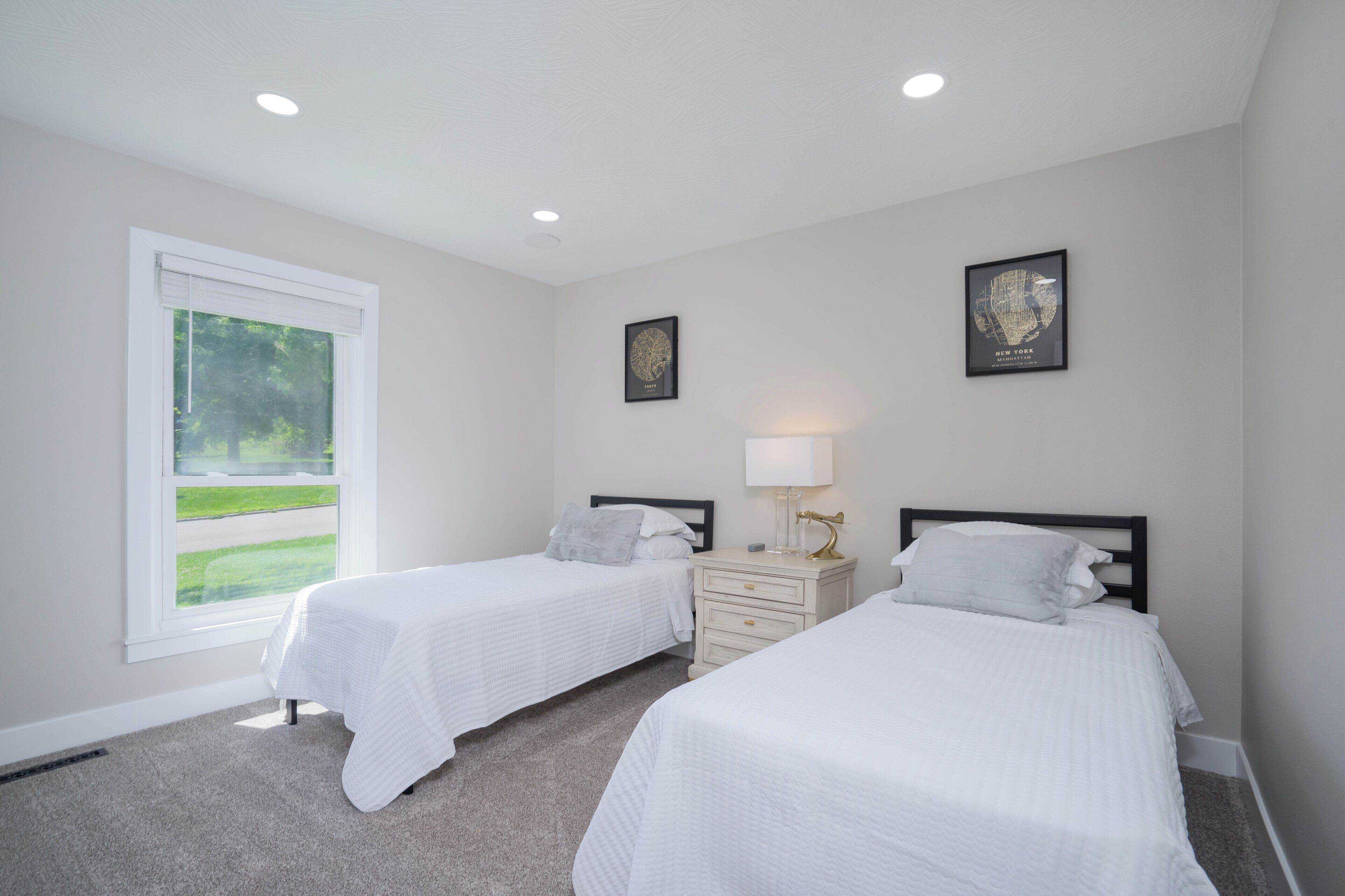 A bedroom with two beds, a nightstand, a lamp, and a window with a view of greenery.