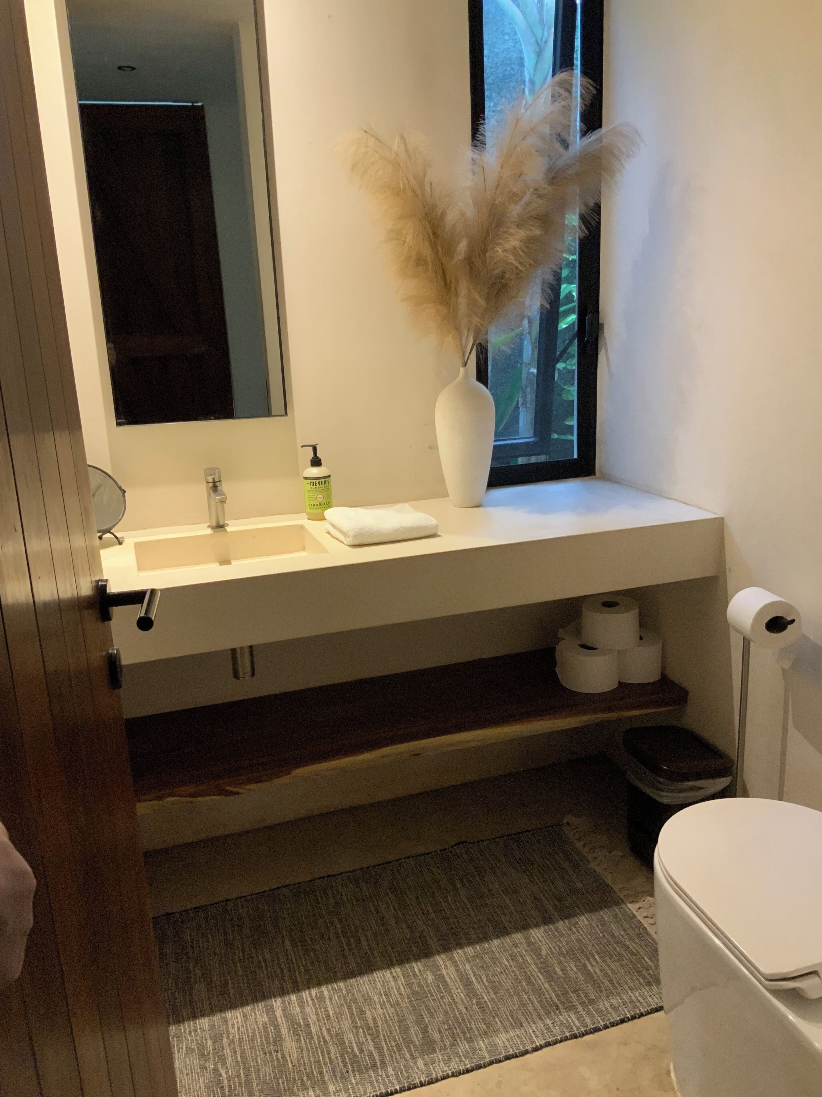 A bathroom with a white vanity, a mirror, a window, a toilet, and a trash bin.