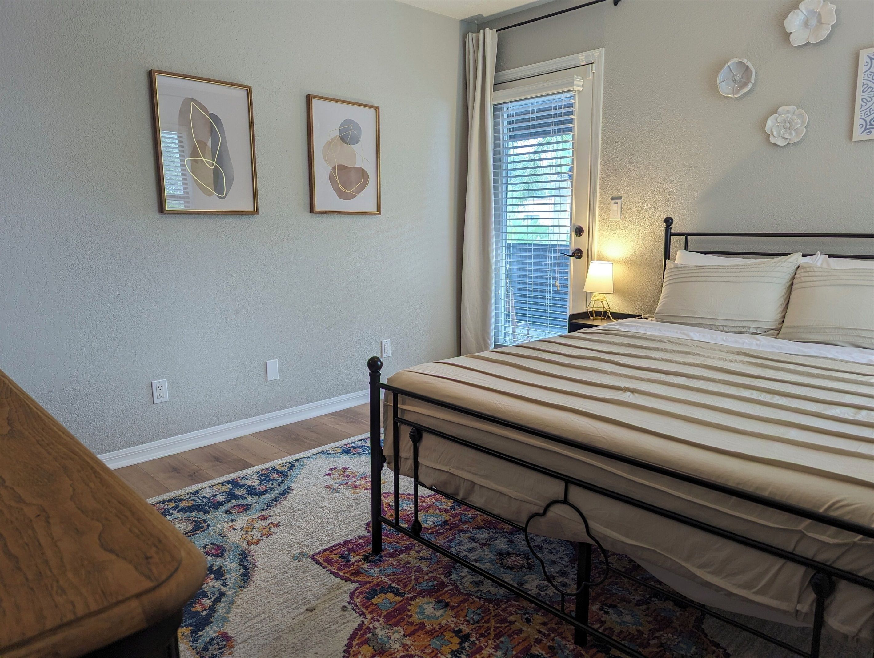 A bedroom with a bed, a nightstand, a lamp, a window with blinds, and two framed abstract artworks on the wall.