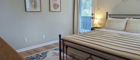A bedroom with a bed, a nightstand, a lamp, a window with blinds, and two framed abstract artworks on the wall.