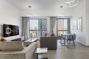 A modern living room with a flat-screen TV, a sofa, a dining table with chairs, and large windows offering a city view. - Beachside Haven Stylish 1 BHK in JBR Dubai (Dubai)