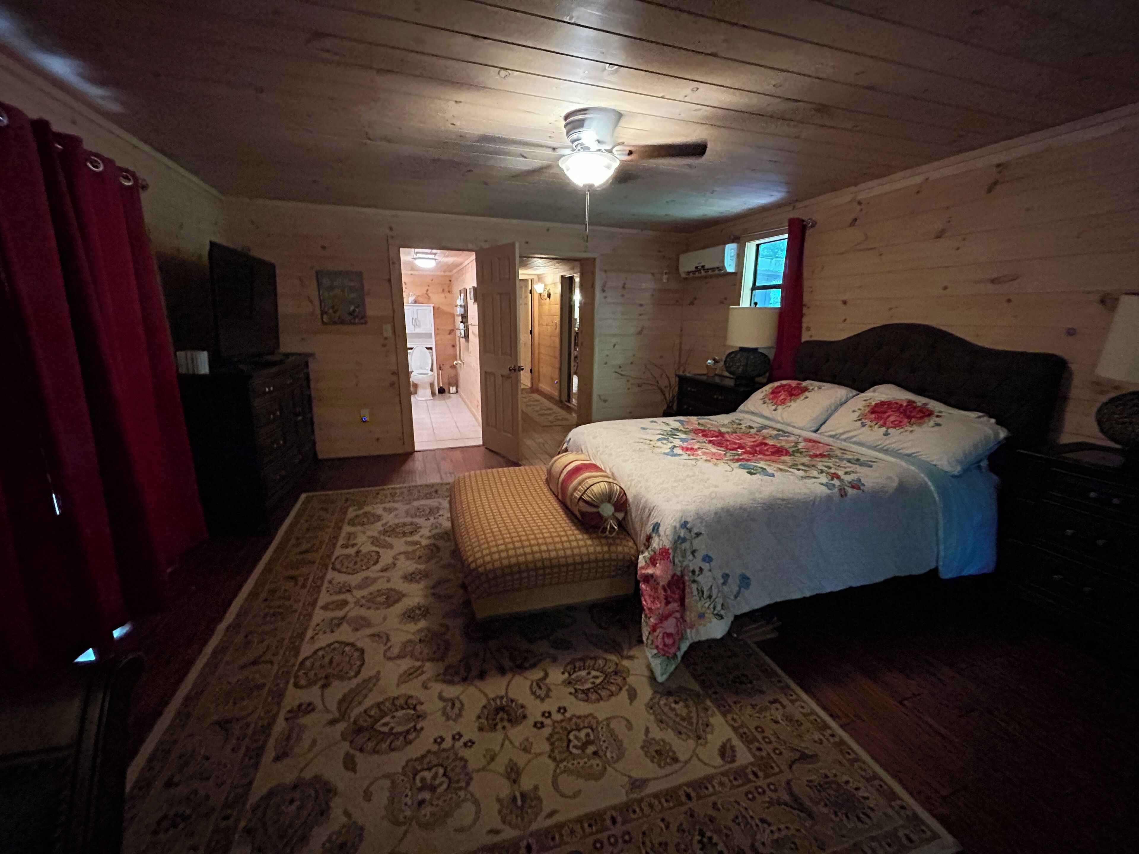 Een slaapkamer met een houten plafond, een bed met bloemenbedrukking, een nachtkastje, een dressoir en een rood gordijn.