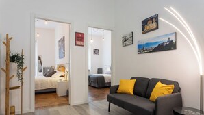 A modern living room with a grey sofa, yellow cushions, a wooden coat rack, and a small table. There are two beds in the background, one with a white duvet and the other with a grey duvet. - Sulla via del treno by Wonderful Italy (Turin)