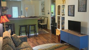 A living room with a sofa, a television on a stand, a kitchen area with a dining table, and a hallway leading to a bedroom. - The Lazy Susan (Charleston)