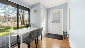 A dining area with a white table and patterned chairs, a large window with a view of a park, and a white door with a glass panel.