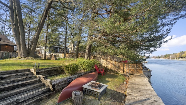 A wooden dock extending into a calm body of water, surrounded by trees and a grassy area with a red canoe and a grill.