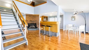 A spacious living area with a wooden staircase, a fireplace, a kitchen area with a bar, and a dining table with chairs.