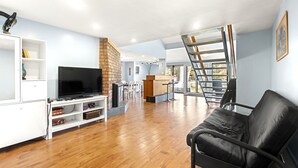 A spacious living area with a flat-screen TV, a black leather sofa, and a wooden floor.