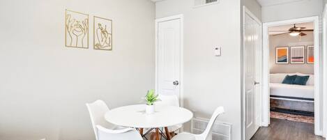 Un coin repas avec une table ronde et des chaises blanches, une plante en pot et deux œuvres d’art encadrées accrochées au mur.