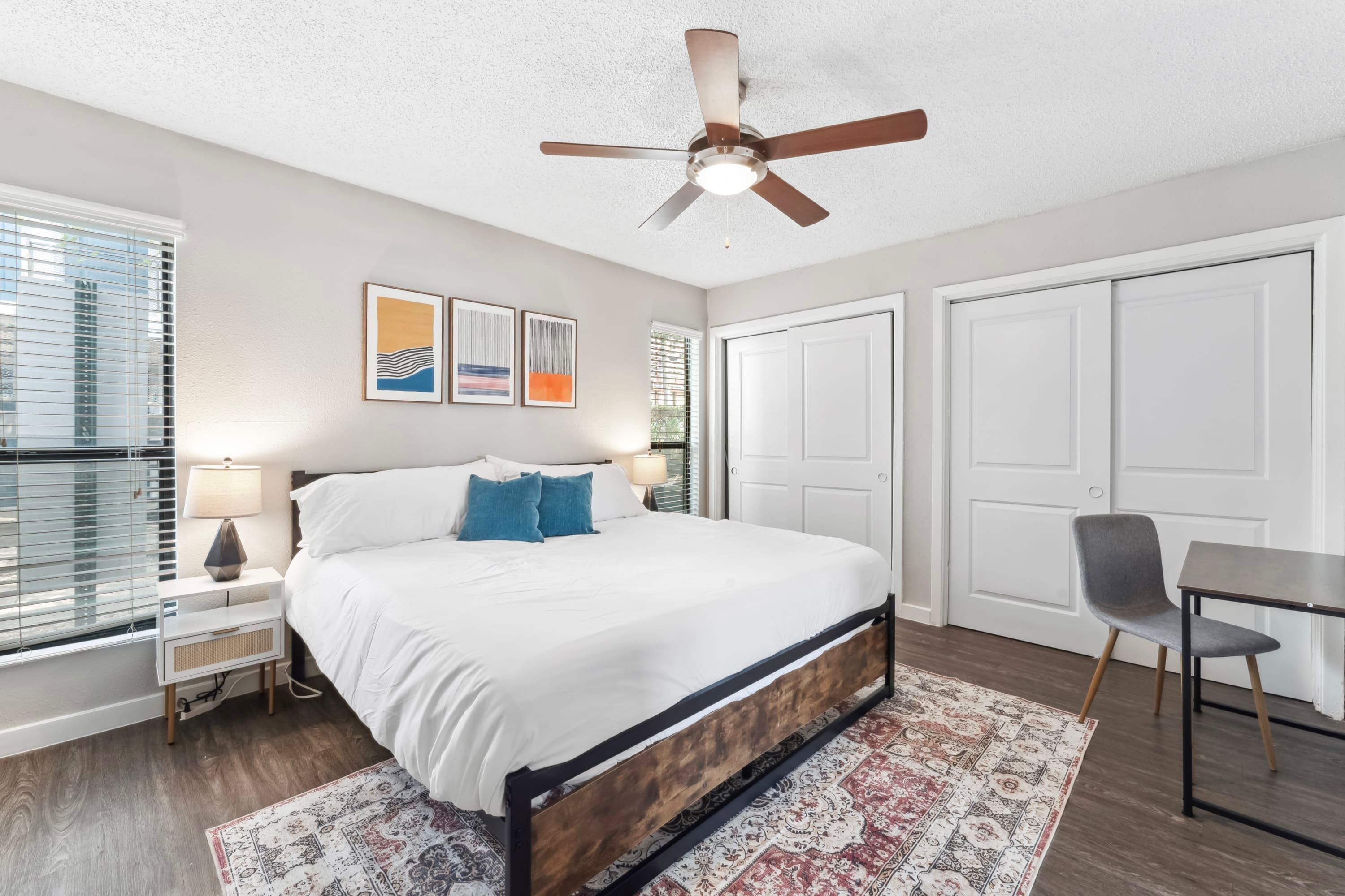 A bedroom with a bed, a ceiling fan, a nightstand with a lamp, a chair, and a table.