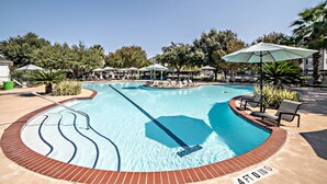 Una gran piscina al aire libre con varias pistas de natación, rodeada de sillones de descanso y sombrillas, y protegida por árboles.