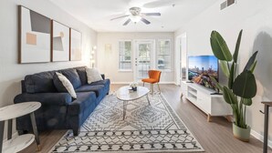 Una sala de estar con un sofá azul, una mesa de centro, un mueble blanco para televisión y una planta grande.