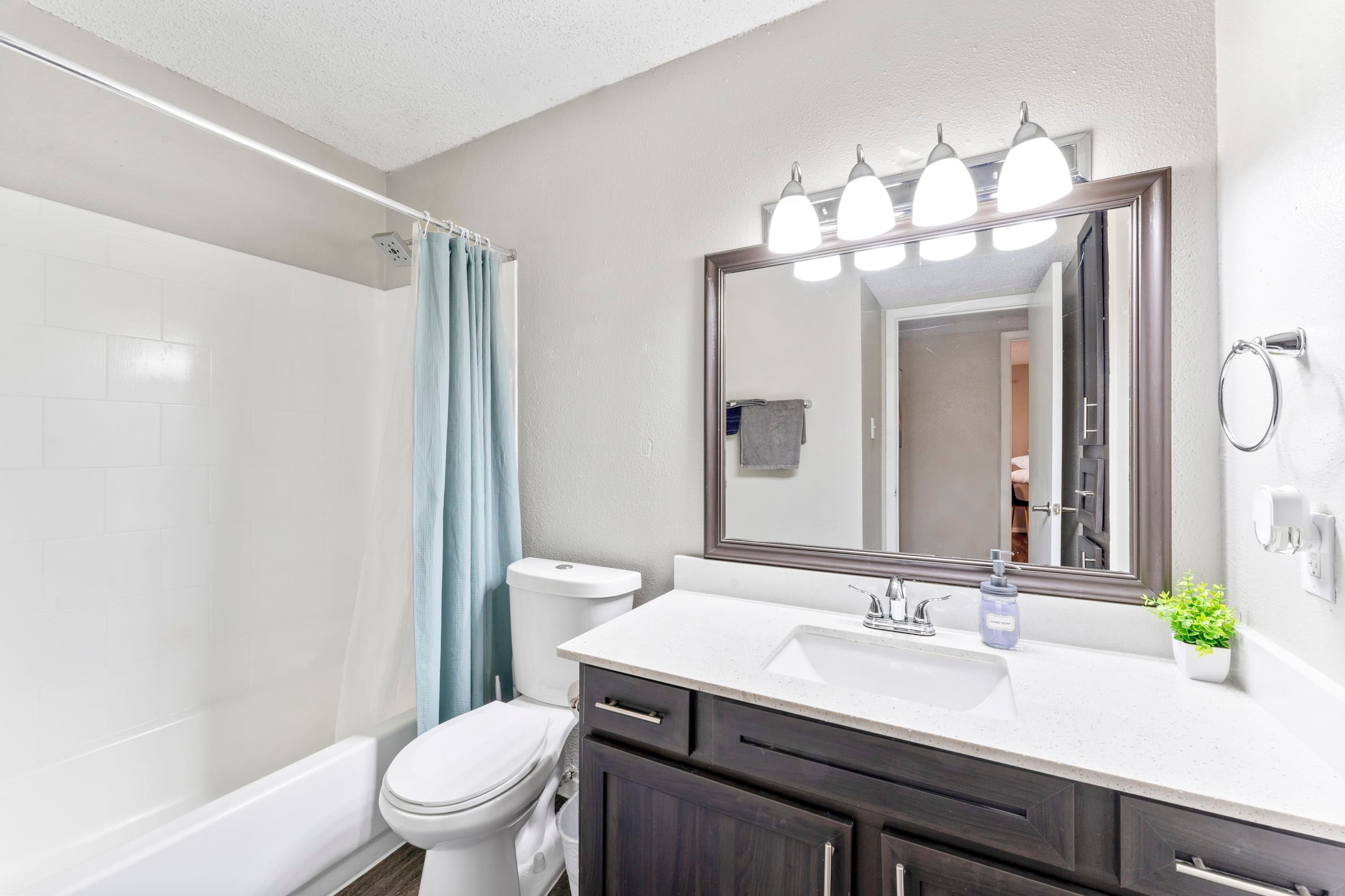 A modern bathroom with a white toilet, a shower with a blue curtain, a large mirror with lights, and a white sink with dark cabinets.