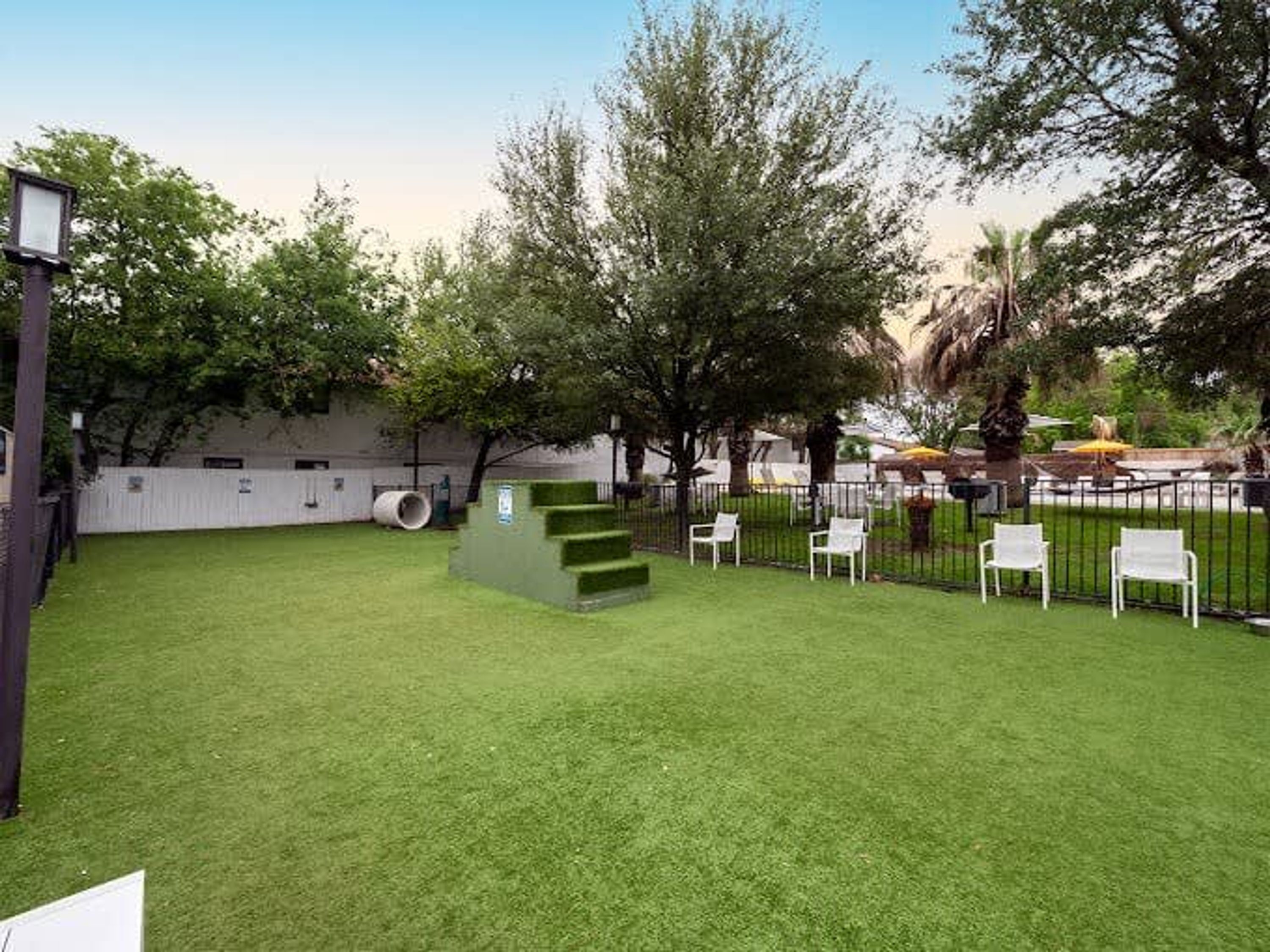 Un área verde bien cuidada con una pequeña escalera, sillas blancas y una valla.