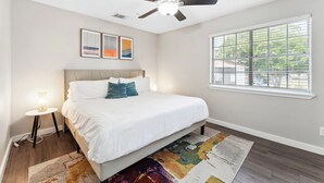 Un dormitorio con cama, ventilador de techo, ventana y una pequeña mesa con lámpara.