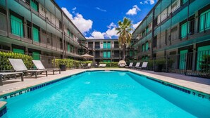 Una piscina con sillones de descanso y una palmera, rodeada por un edificio de varios pisos con balcones.