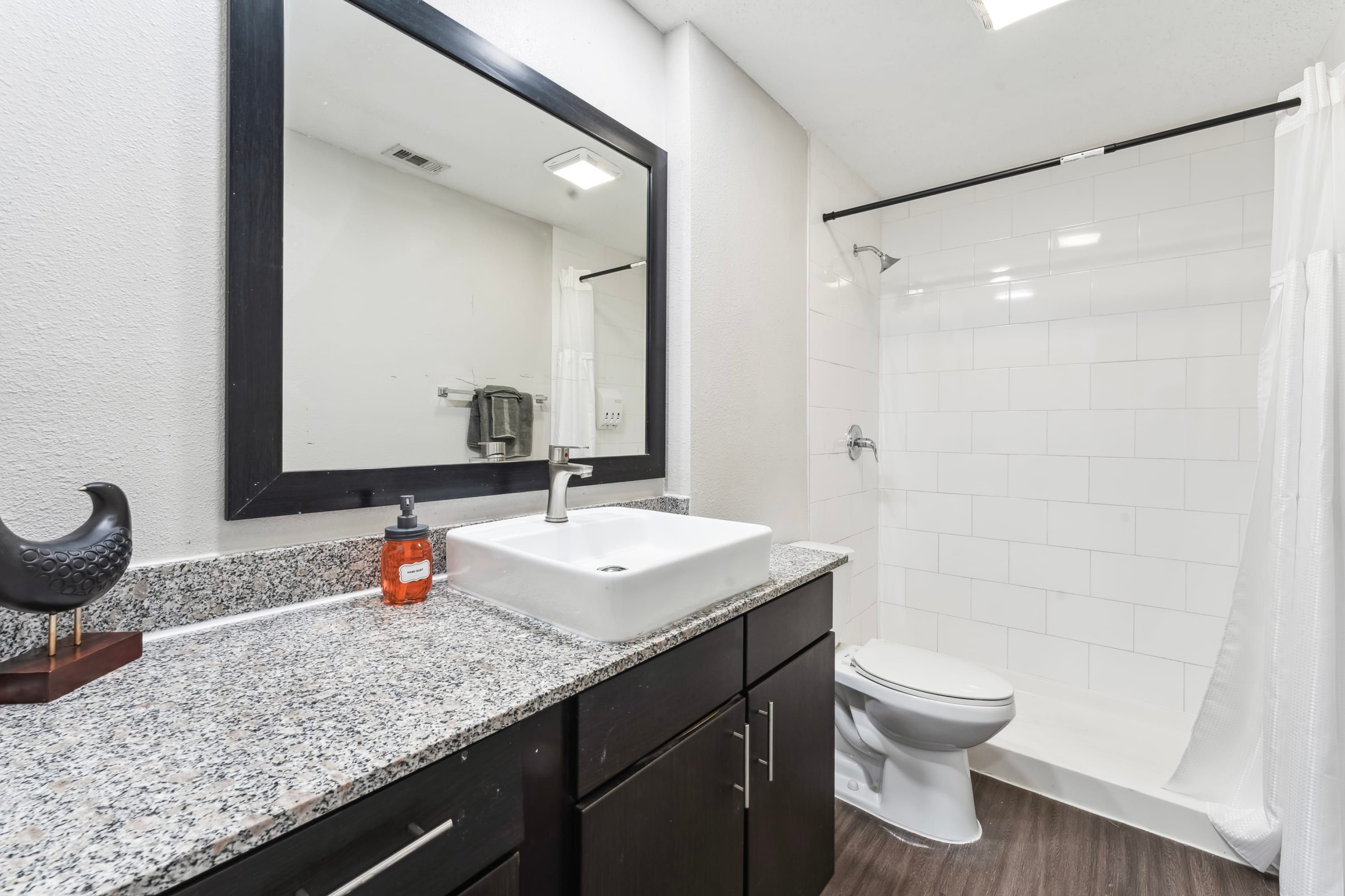 Un baño moderno con encimera de granito, un lavabo blanco, un armario negro, un inodoro y una cabina de ducha con mampara de vidrio.