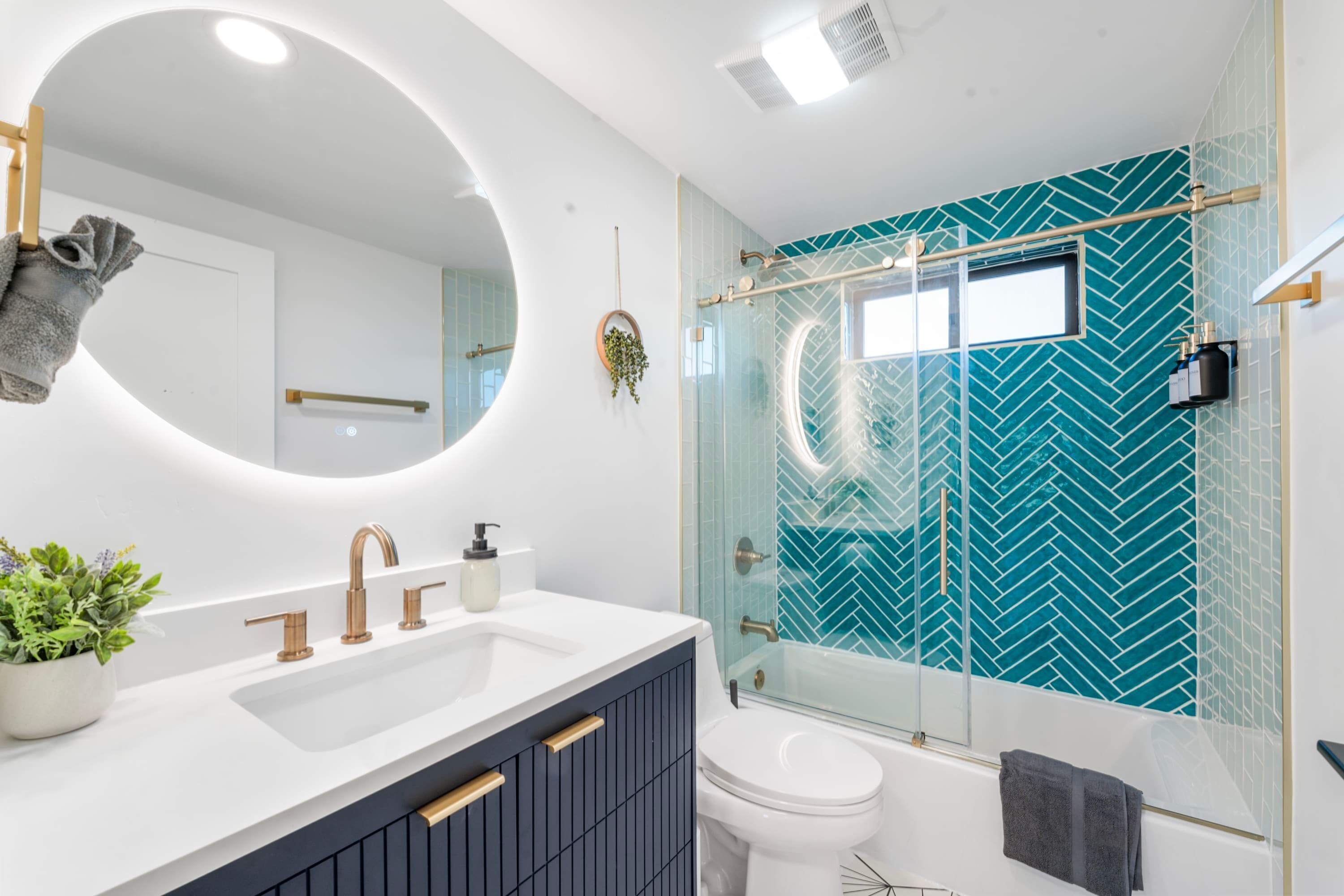 A modern bathroom with a white sink, a round mirror, a glass shower enclosure, and a teal patterned wall.