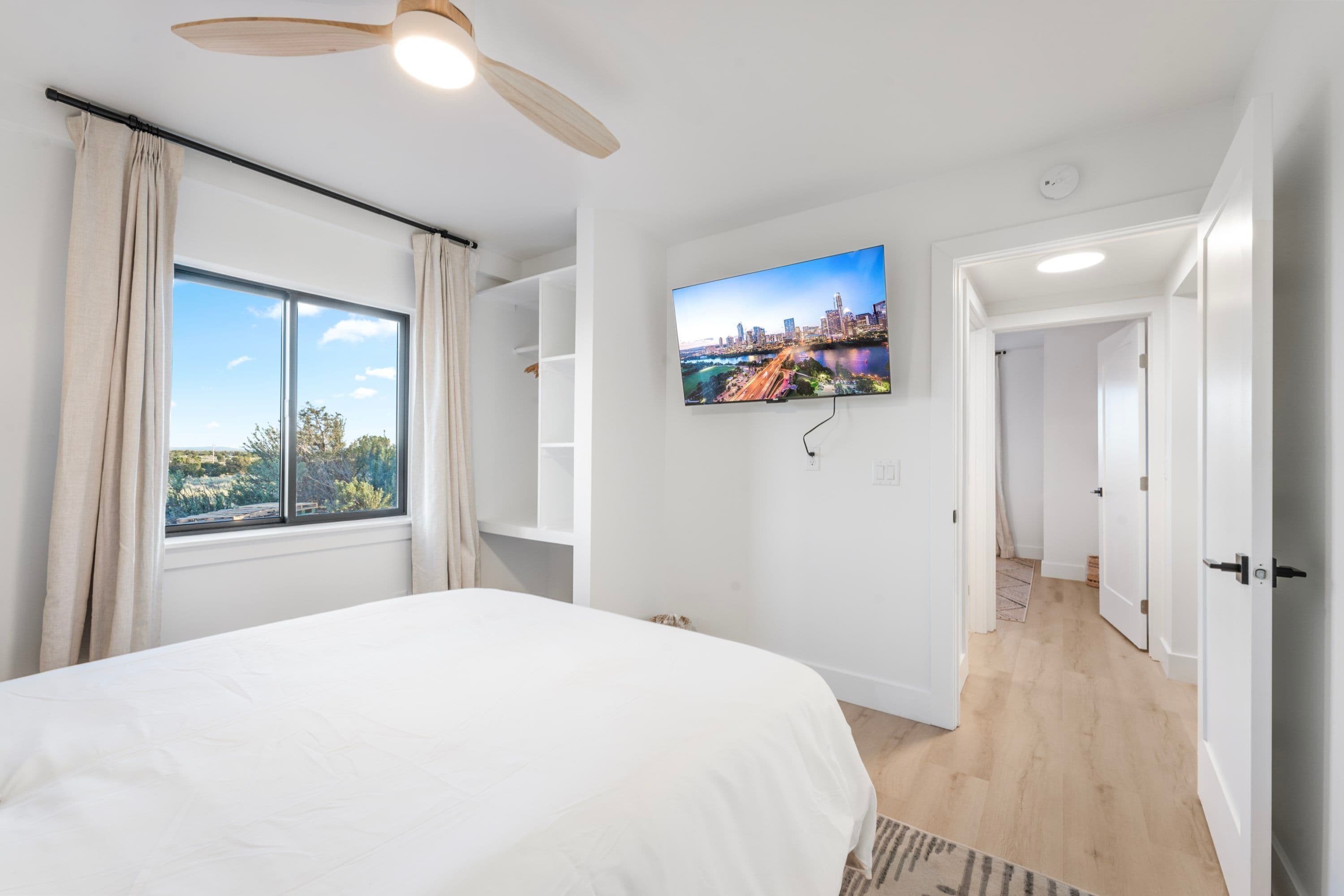 A bedroom with a large bed, a ceiling fan, a TV mounted on the wall, and a window with curtains.