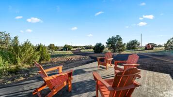 A deck with wooden chairs and a table, surrounded by greenery and a clear sky.