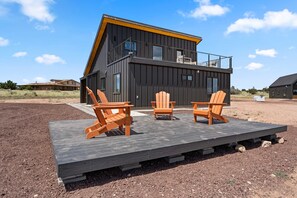 A modern two-story house with a balcony, a deck with three wooden chairs, and a clear sky. - NEW Luxe Desert Retreat Near Grandcanyon S Rim (Williams)