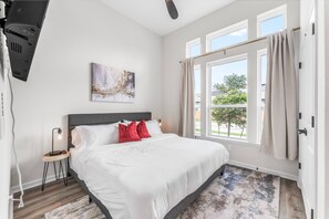 A bedroom with a bed, a nightstand, a lamp, a painting, and a window with curtains.