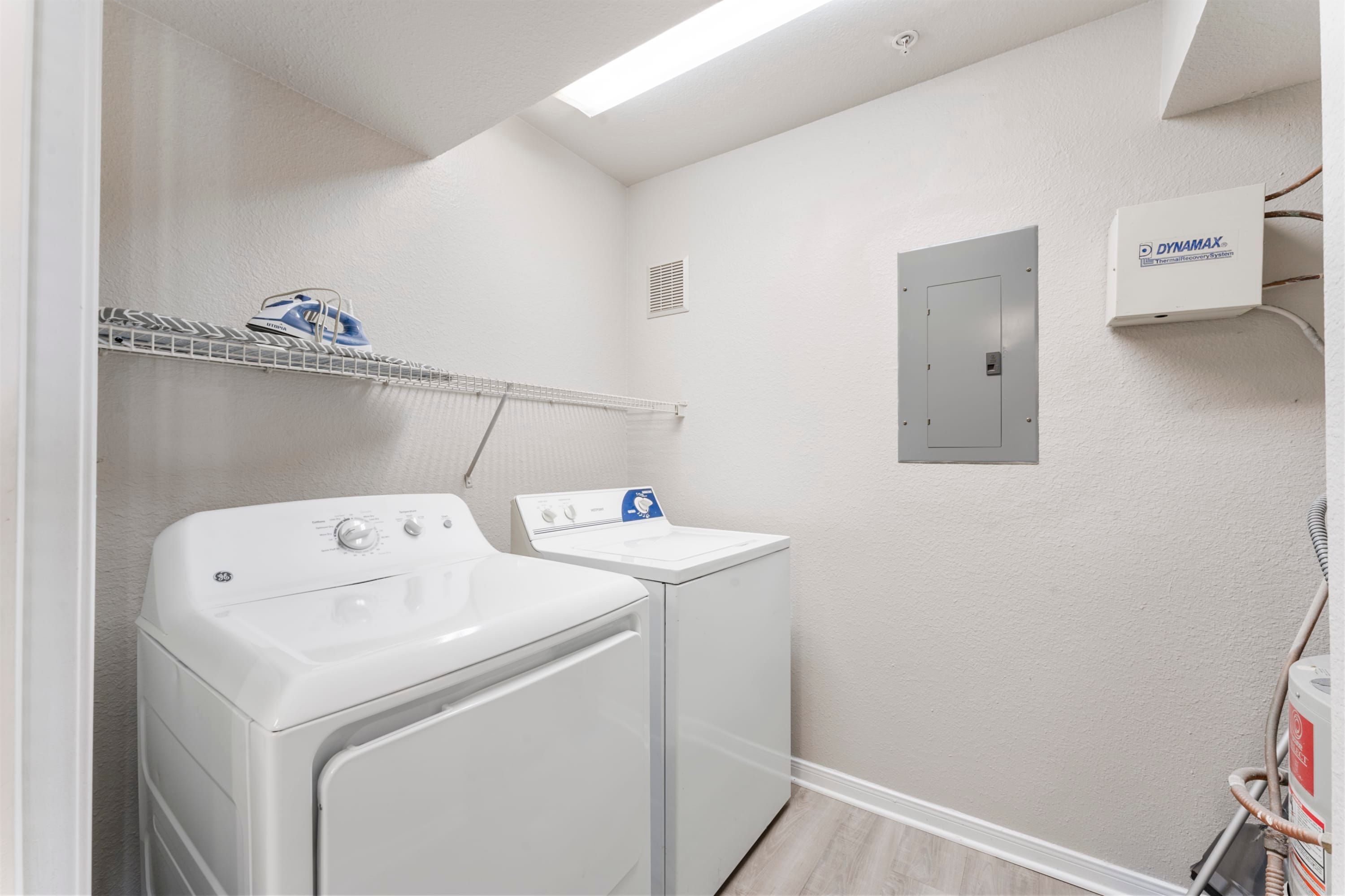A laundry area with two white washing machines, a shelf with a pair of shoes, a wall-mounted air conditioning unit, and a vent.
