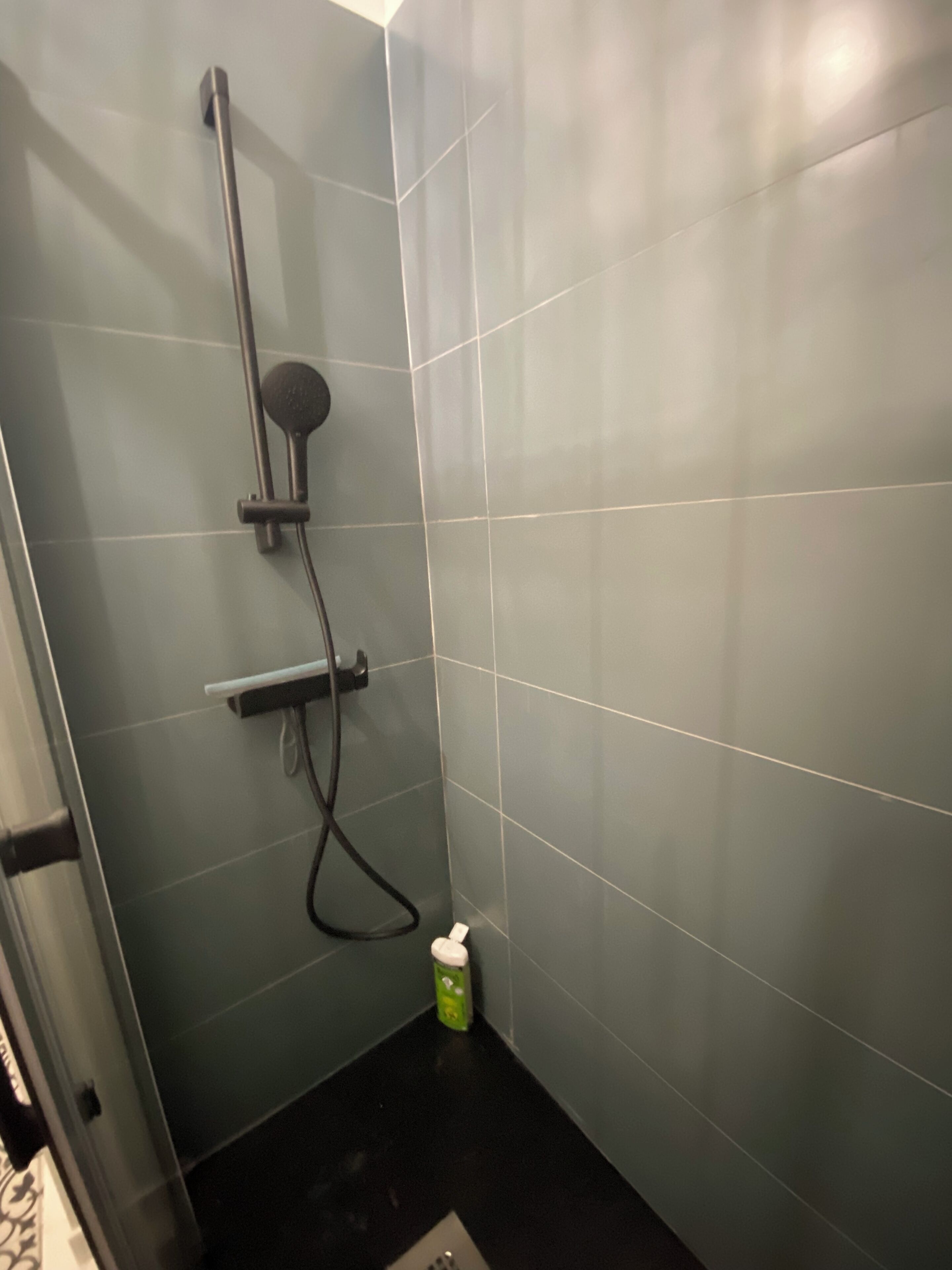 Een klein, afgesloten douchegedeelte met een handdouche, een douchetuitstuk en een fles shampoo op de vloer.