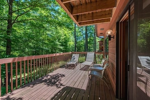 Um deck de madeira com duas espreguiçadeiras e uma mesa, cercado por guarda-corpo de madeira, com vista para uma área arborizada.
