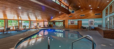 An indoor swimming pool with wooden ceiling beams, clear glass walls, and a tiled floor.