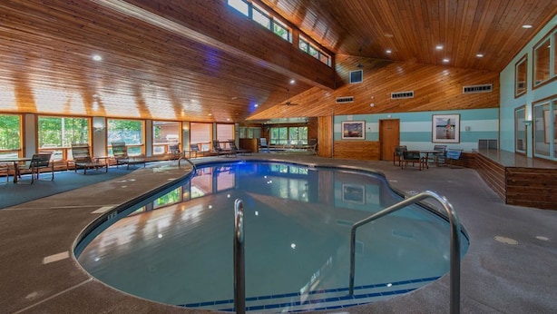 Une piscine intérieure dotée de poutres en bois au plafond, de murs en verre transparent et d’un sol carrelé.