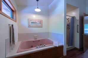 A modern bathroom with a large pink bathtub, a shower with a grab bar, a framed picture on the wall, and a window providing natural light. - 2BR Condo with Fireplace, Balcony, & Lakefront Access at The Rushes (Baileys Harbor)